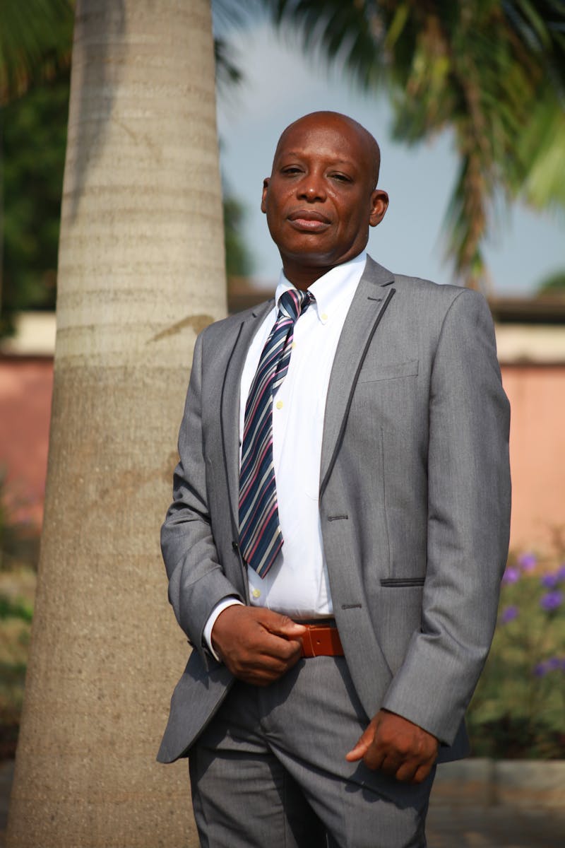 Confident businessman posing in a gray suit with a tie, standing outdoors by a palm tree.