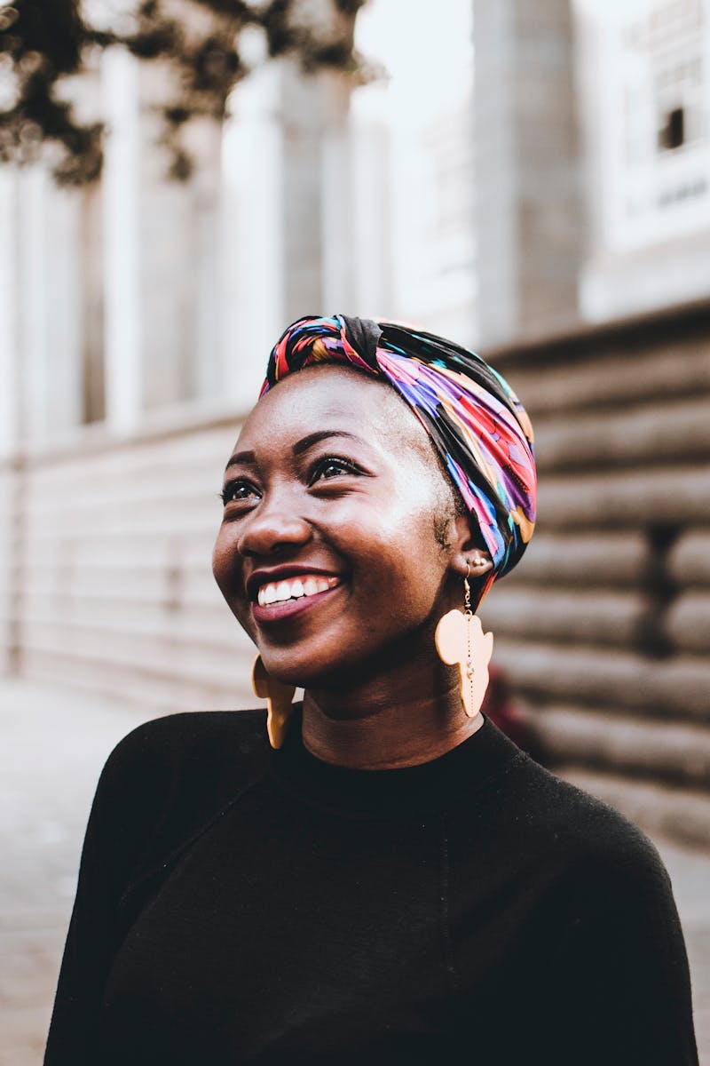 Smiling woman wearing vibrant headscarf and earrings, captured in an urban street setting.